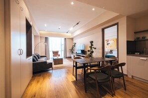 A modern living room with a sofa, dining table, and chairs, a kitchen area with cabinets and a countertop, and a large window providing natural light. - May Seven Hotel and Suites (Ulaanbaatar)
