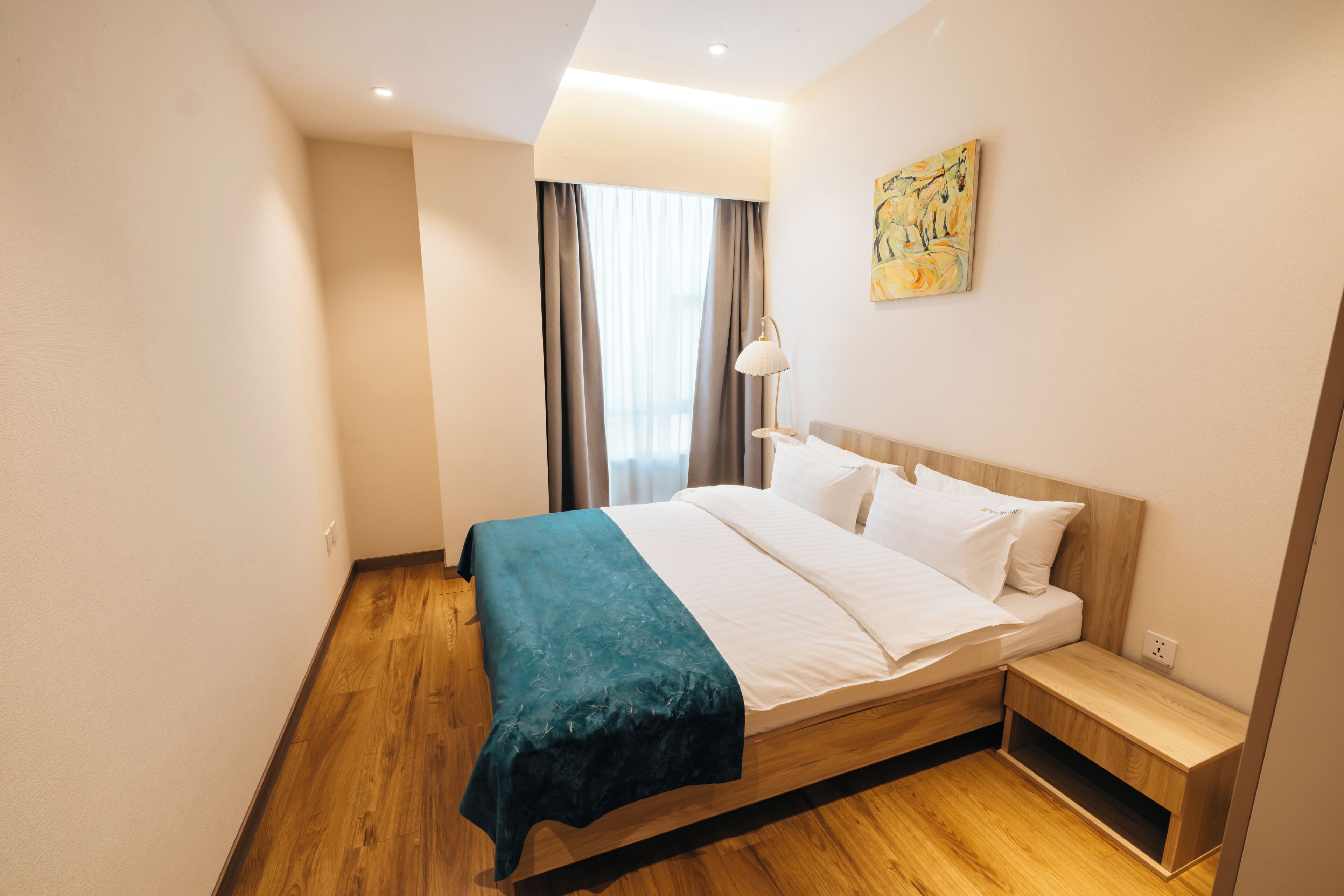 A modern hotel room with a wooden floor, a bed with white linens, a bedside table, and a painting on the wall.