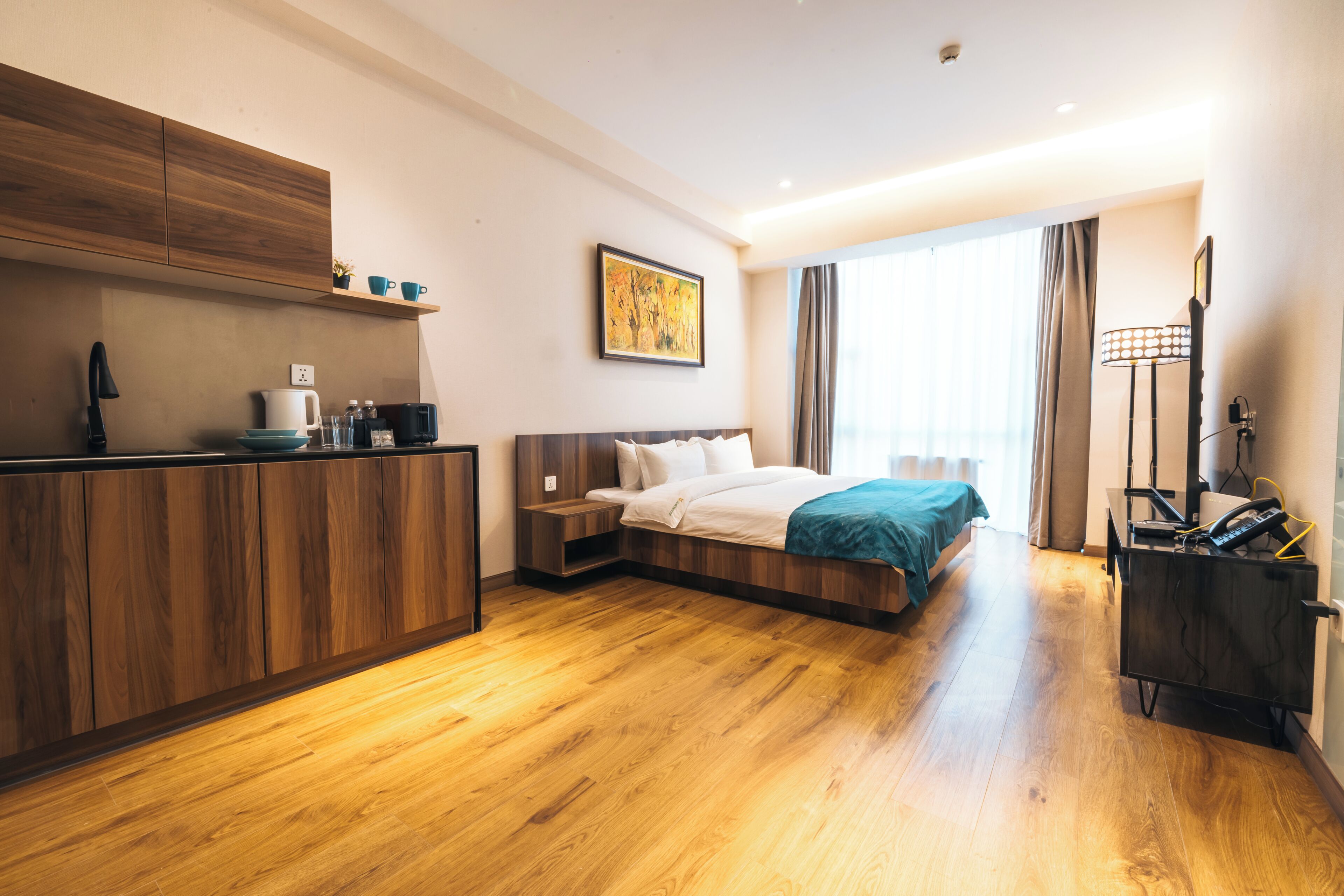 A hotel room with a wooden floor, a bed with a blue blanket, a desk with a lamp, and a painting on the wall.