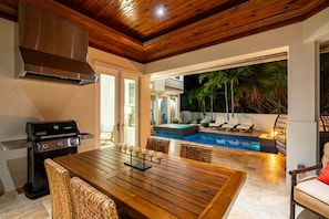 A wooden dining table with candles, a grill, and wicker chairs in a patio area with a pool and lounge chairs. - Upscale 6BR Mansion w/ Pool & Hot Tub (Stuart)