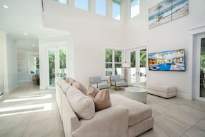 A spacious living room with a large beige sofa, a round coffee table, and a flat-screen TV mounted on the wall. - Upscale 6BR Mansion w/ Pool & Hot Tub (Stuart)