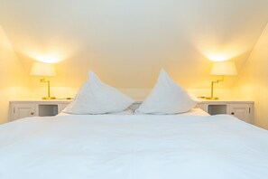 Ein ordentlich bezogenes Bett mit weißen Leinen und zwei großen weißen Kissen. An beiden Seiten des Bettes stehen Nachttische mit Lampen.