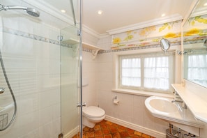 A bathroom with a glass-enclosed shower, a toilet, a sink, and a window with floral wallpaper.