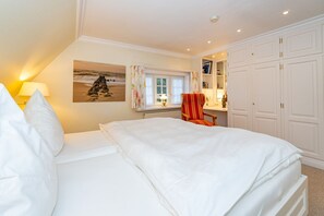 Ein Schlafzimmer mit einem Bett, einem Sessel, einem Fenster mit Vorhängen, einem Bild an der Wand und einem Kleiderschrank.