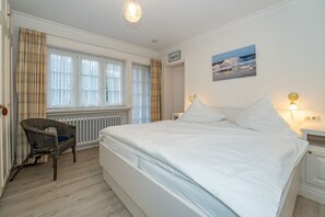 Un dormitorio con una cama grande, una silla, una ventana con cortinas, un cuadro en la pared y una lámpara de noche.