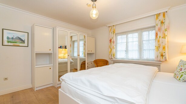 Ein Schlafzimmer mit einem großen Bett, einem Schrank mit Spiegel, einem Sessel und einem Fenster mit Vorhängen.