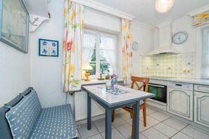 A kitchen with a dining table, chairs, a radiator, a clock, and a window with curtains. - Kajüte - Friesische 2-zimmerwohnung mit Terrasse (Sylt)