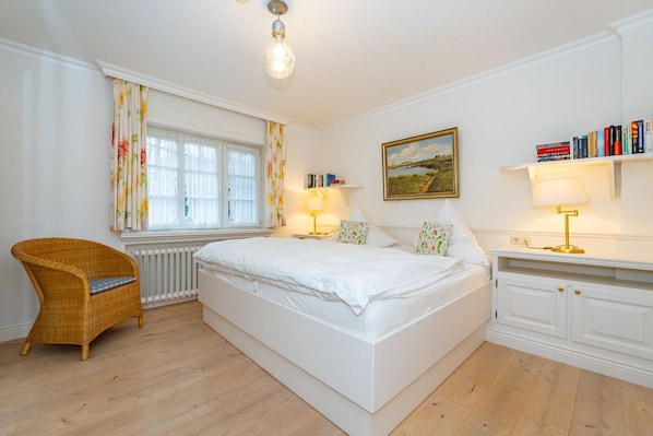 A bedroom with a large bed, a wicker chair, a radiator, a bookshelf, and a painting on the wall.