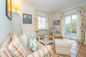 A cozy living room with a sofa, a small table, and a chair. There is a window with curtains and a view of the outside. - Kajüte - Friesische 2-zimmerwohnung mit Terrasse (Sylt)
