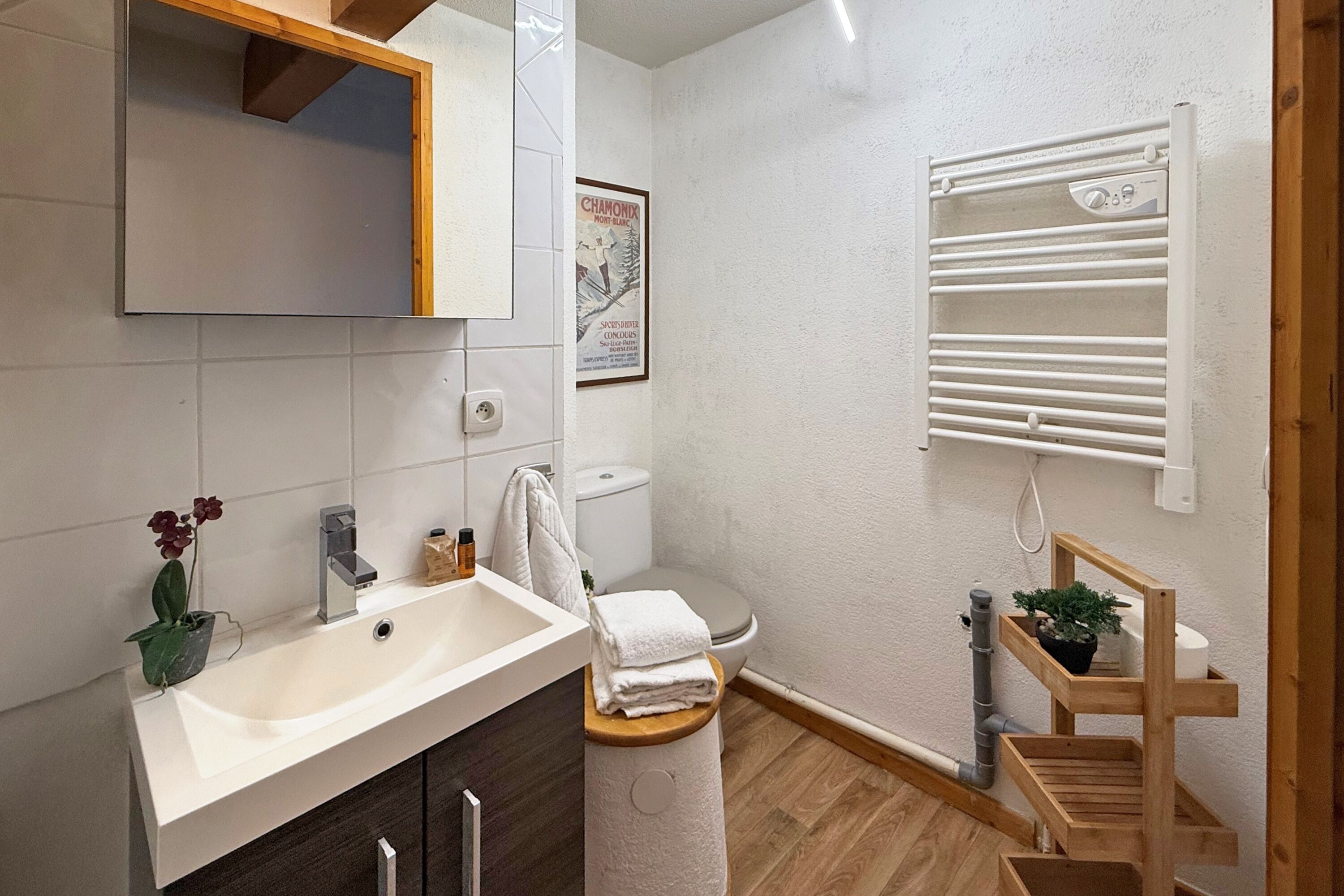 Un bagno moderno con lavandino, specchio e vaso sospeso bianchi. Una mensola in legno con una pianta e un asciugamano.