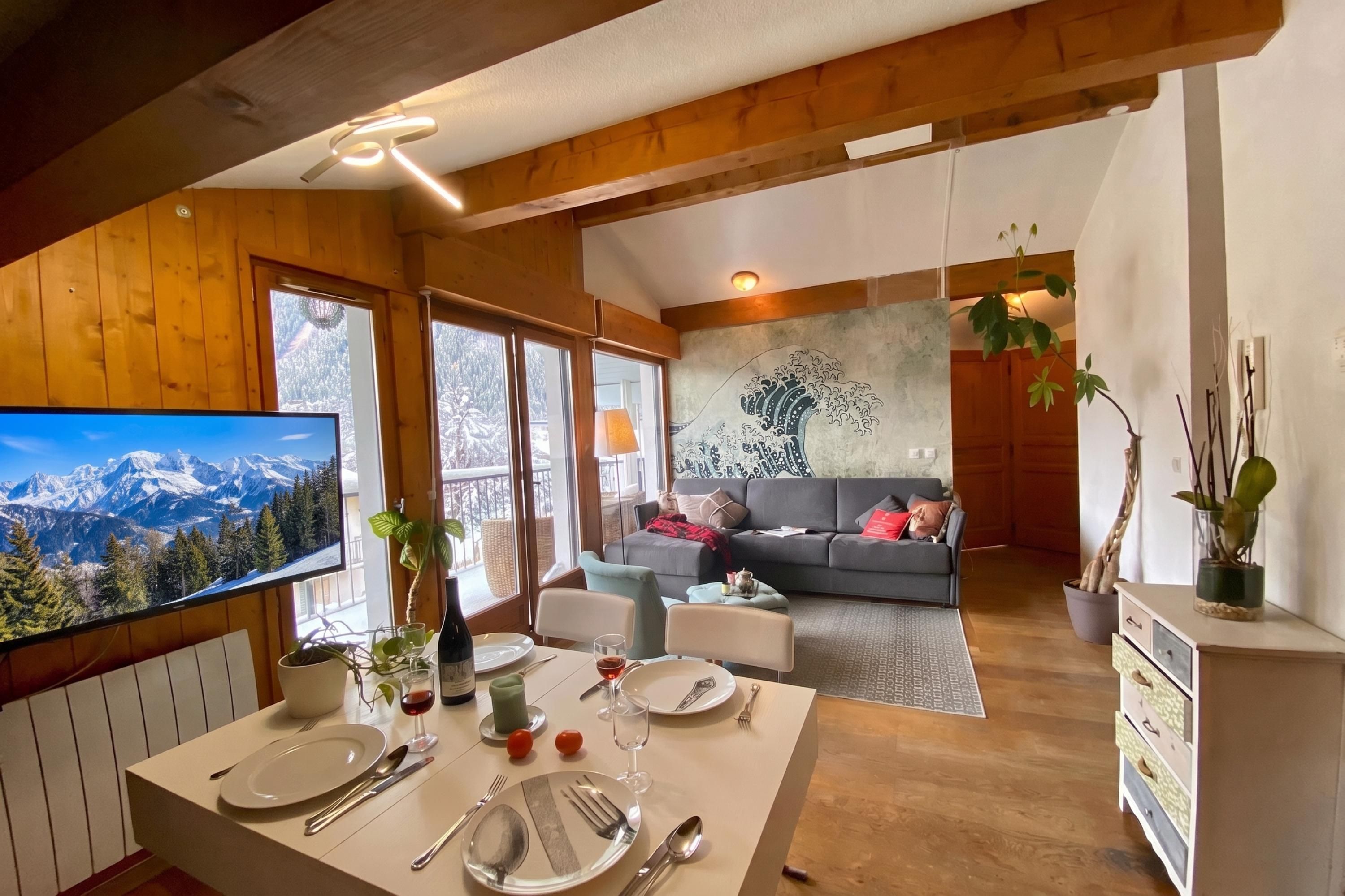 Un coin repas avec une table dressée pour deux personnes, un canapé en arrière-plan et un grand téléviseur diffusant une vue sur la montagne.
