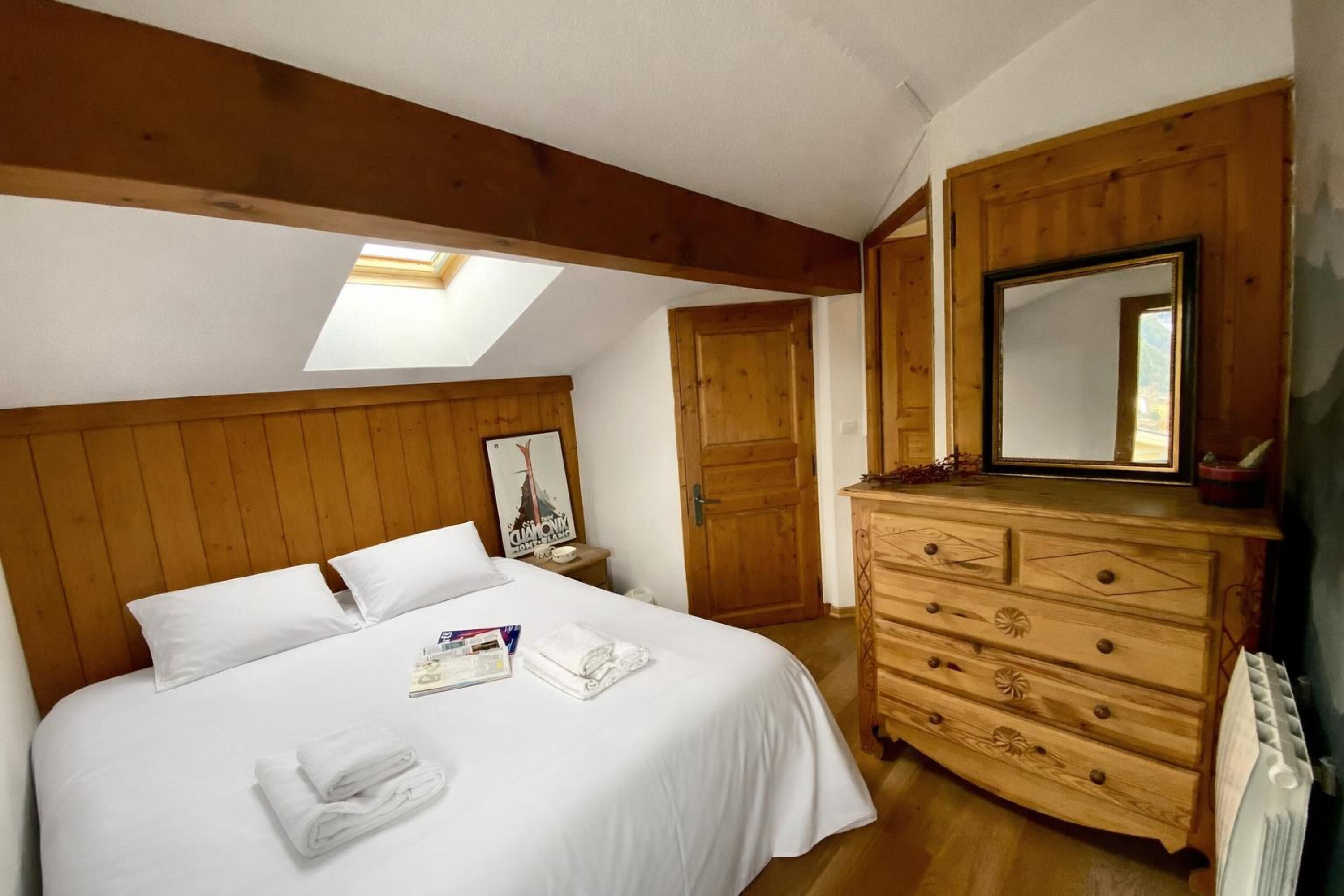 Un dormitorio con cabecera de madera, cama con ropa blanca, cómoda, espejo y tragaluz.