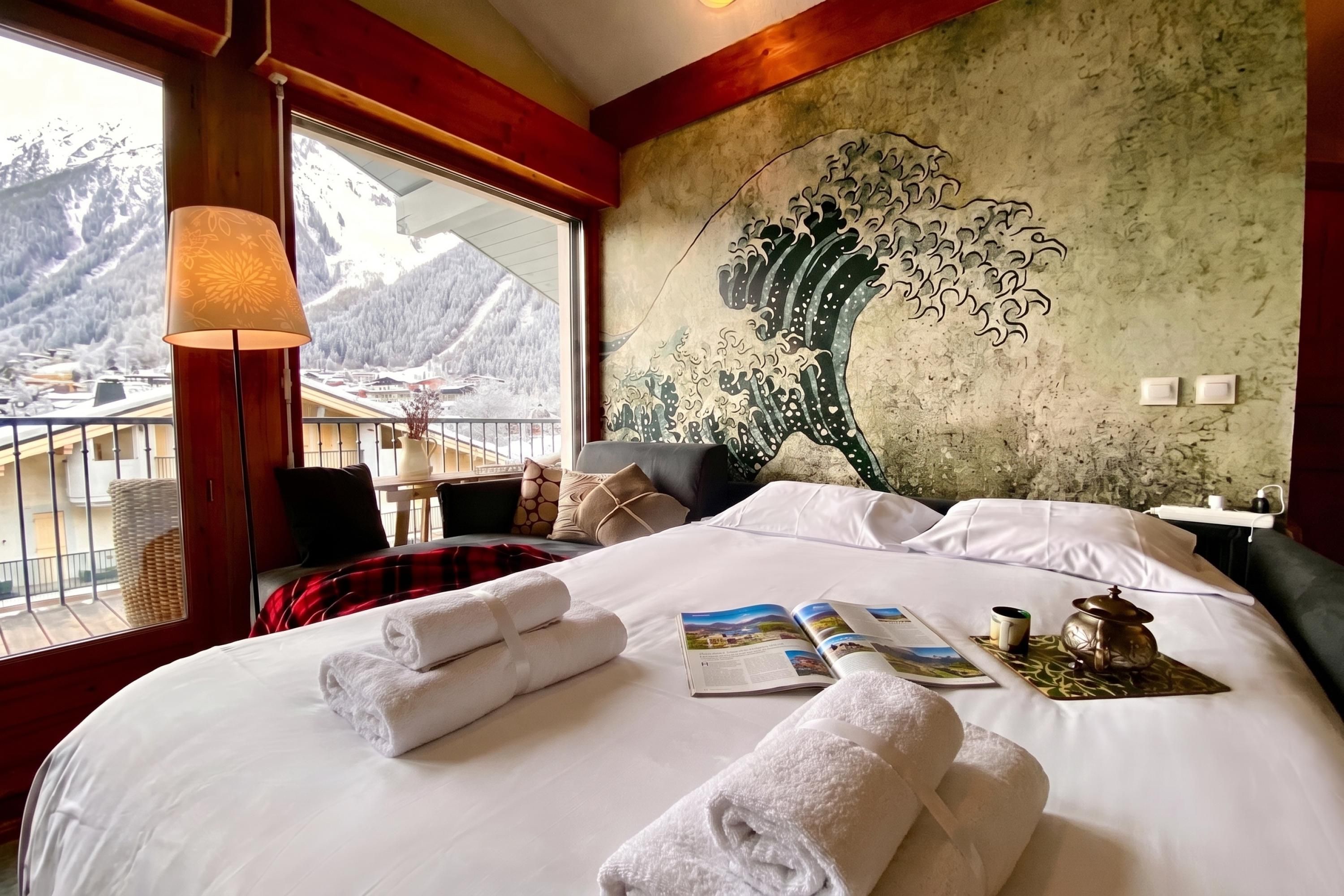 A hotel room with a bed, a view of mountains, a wall with a wave painting, and a lamp.