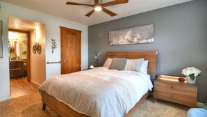 A bedroom with a wooden bed, a nightstand with a lamp, a ceiling fan, and a painting on the wall.
