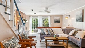 A spacious living room with a large sofa, a coffee table, and wooden chairs. There is a staircase with rope handrails on the left. - Steps to Beach | Backyard | Shuffle Board (Hampton)