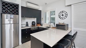 A modern kitchen with a stainless steel refrigerator, a microwave, a toaster, and a coffee maker. There is a clock on the wall and a window with blinds. - 1 Bedroom Apartment Beach Club Palm Cove (Palm Cove)