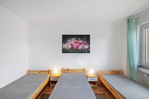 A small room with two single beds, a wooden headboard, bedside tables with lamps, a painting on the wall, and a window with curtains.