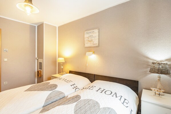 A neatly made bed with a 'HOME HOME HOME' pillow, a wall-mounted light, and a small decorative item on the nightstand.