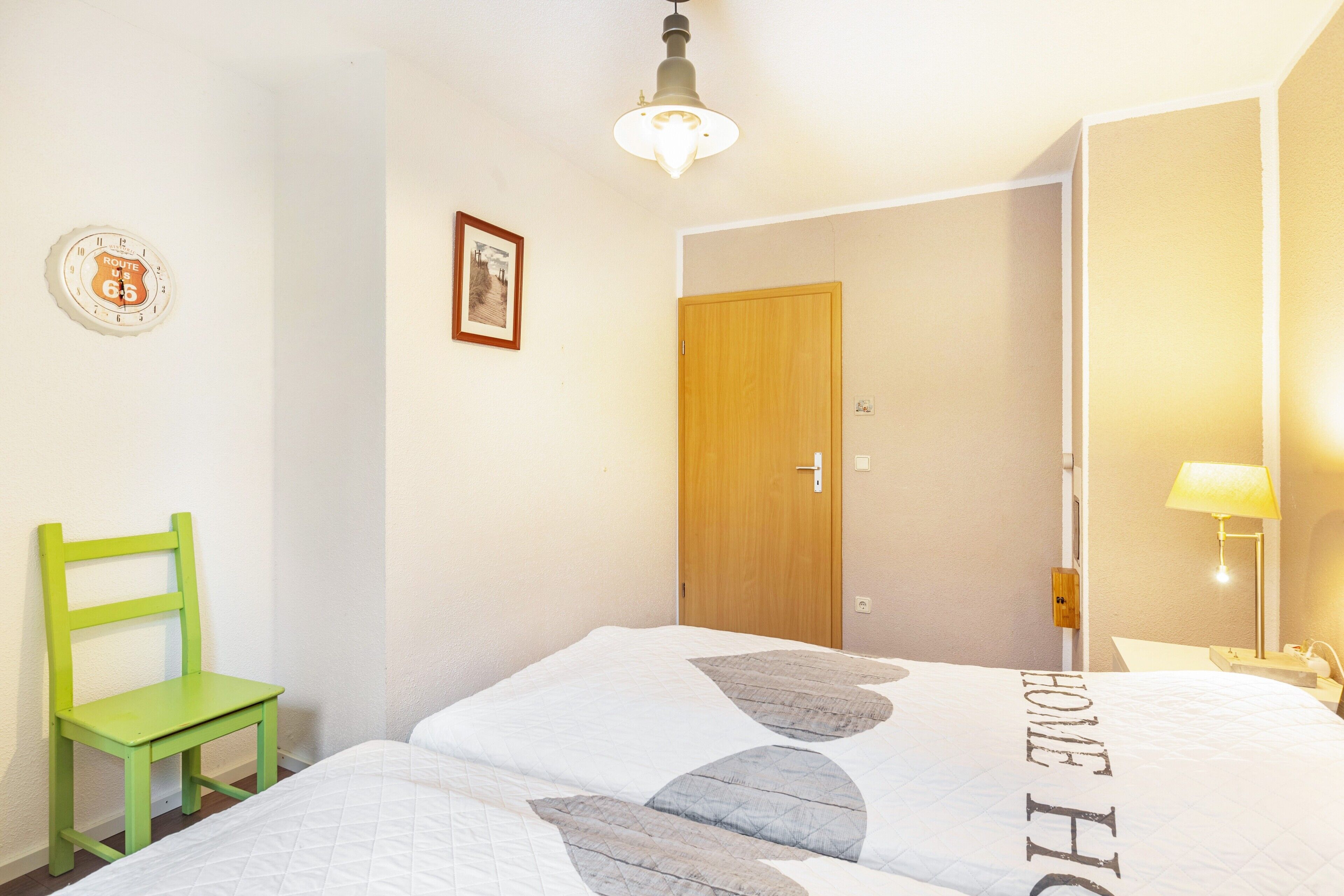 Een kamer met een bed, een stoel, een deur, een wandlamp en een kleine tafel met een lamp.