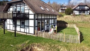 Perinteinen puurunkoineen osittain puulla verhoiltu talo, jossa on parveke, aidattu piha ja kirkas taivas.