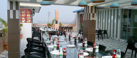 An outdoor dining area with tables set for two, black chairs, and blue pendant lights.
