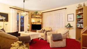 A living room with a red carpet, beige armchairs, a table with a vase of flowers, a bookshelf, and a television. - Apartment 'Michael' with Balcony and Wi-Fi (Langenargen)