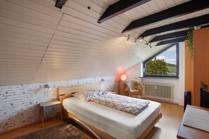 Une chambre avec un plafond en pente, un lit avec une literie à motifs, une table de chevet, une chaise et une vue sur l’extérieur par une fenêtre.