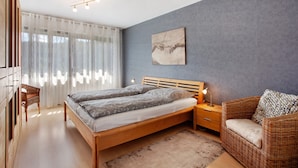 A bedroom with a wooden bed, a wicker chair, a nightstand, and a large window with curtains. - Apartment 'Lux' with Private Garden, Balcony and Wi-Fi (Plettenberg)