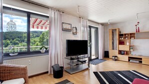 A modern living room with a large window, a flat-screen TV, a wicker sofa, a wooden cabinet, and a view of a forest. - Apartment 'Lux' with Private Garden, Balcony and Wi-Fi (Plettenberg)