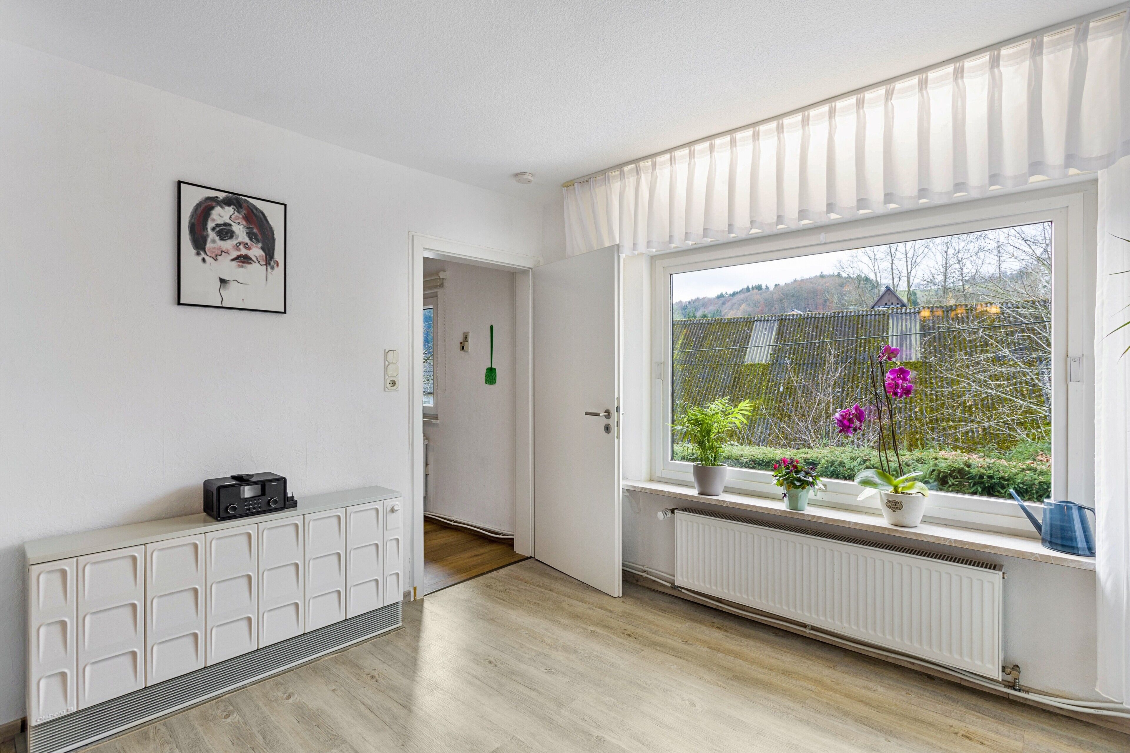 Una habitación con radiador, un armario, una ventana con plantas y una puerta que da a otra estancia.