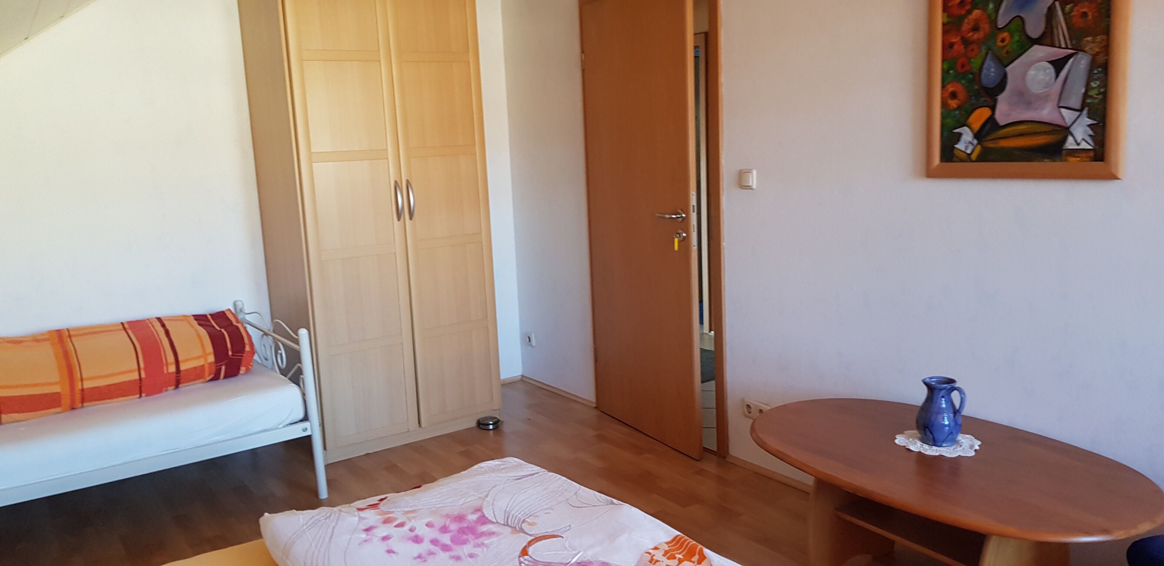 Een kamer met een houten garderobe, een witte bank, een klein tafeltje met een blauwe kruik, en een schilderij aan de muur.