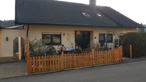 Uma casa de dois andares com telhado de telhas, cerca de madeira e jardim frontal.