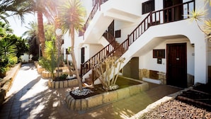 A two-story building with a white facade, a wooden staircase, and a small garden area with palm trees and plants. - Paraiso Royal I, Sunny Terrace & Heated Pool (Playa de las Américas)