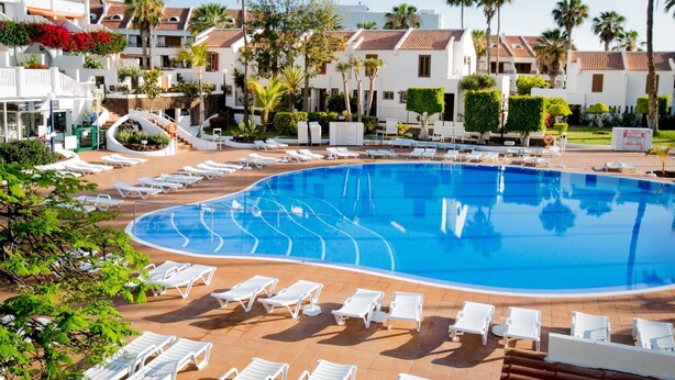 A large swimming pool with white lounge chairs arranged around it, surrounded by palm trees and buildings with tiled roofs. - 395 Lovely Duplex Parque Santiago II (Playa de las Américas)