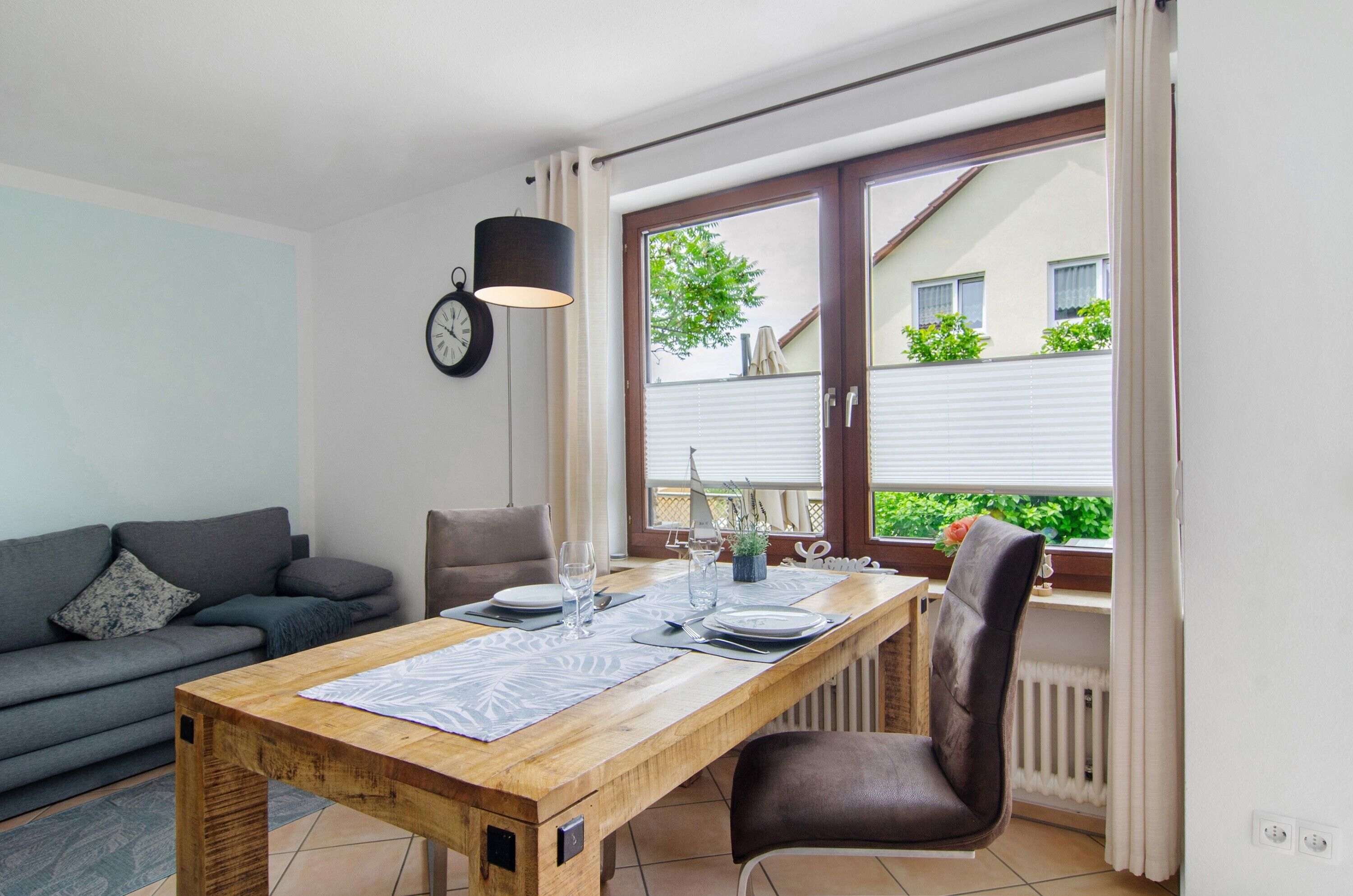 Ein Essbereich mit einem Holztisch, gedeckt für vier Personen, einem grauen Sofa, einer Uhr und einem Fenster mit Jalousien.