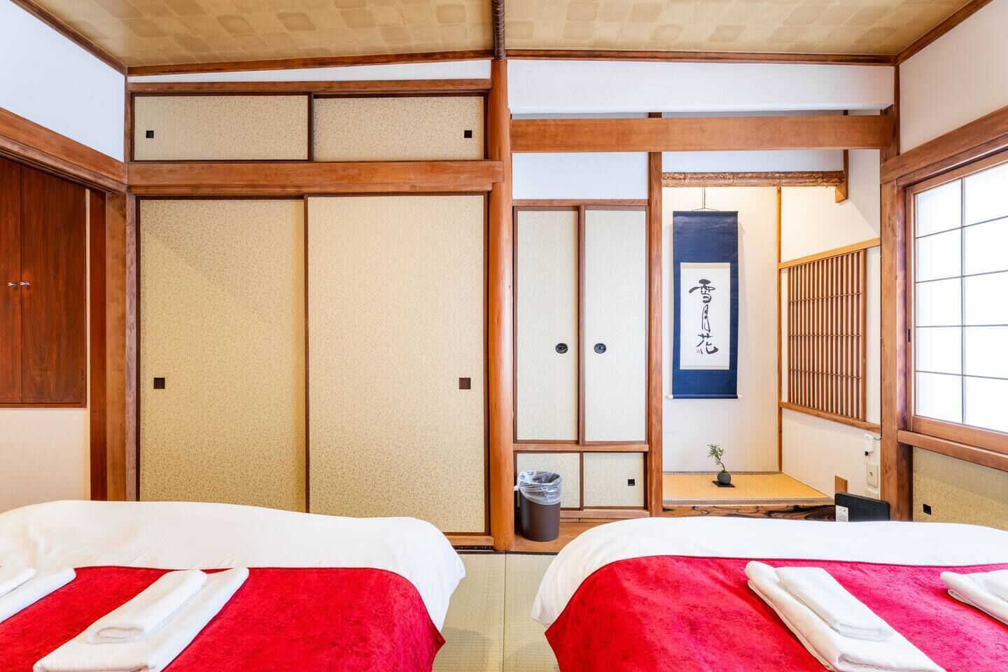 A traditional Japanese room with tatami flooring, two beds with red and white bedding, a shoji screen, and a hanging scroll with calligraphy.