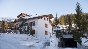 Ein zweistöckiges Chalet mit Holzbalkon, umgeben von schneebedeckten Bäumen und einem Berg im Hintergrund.