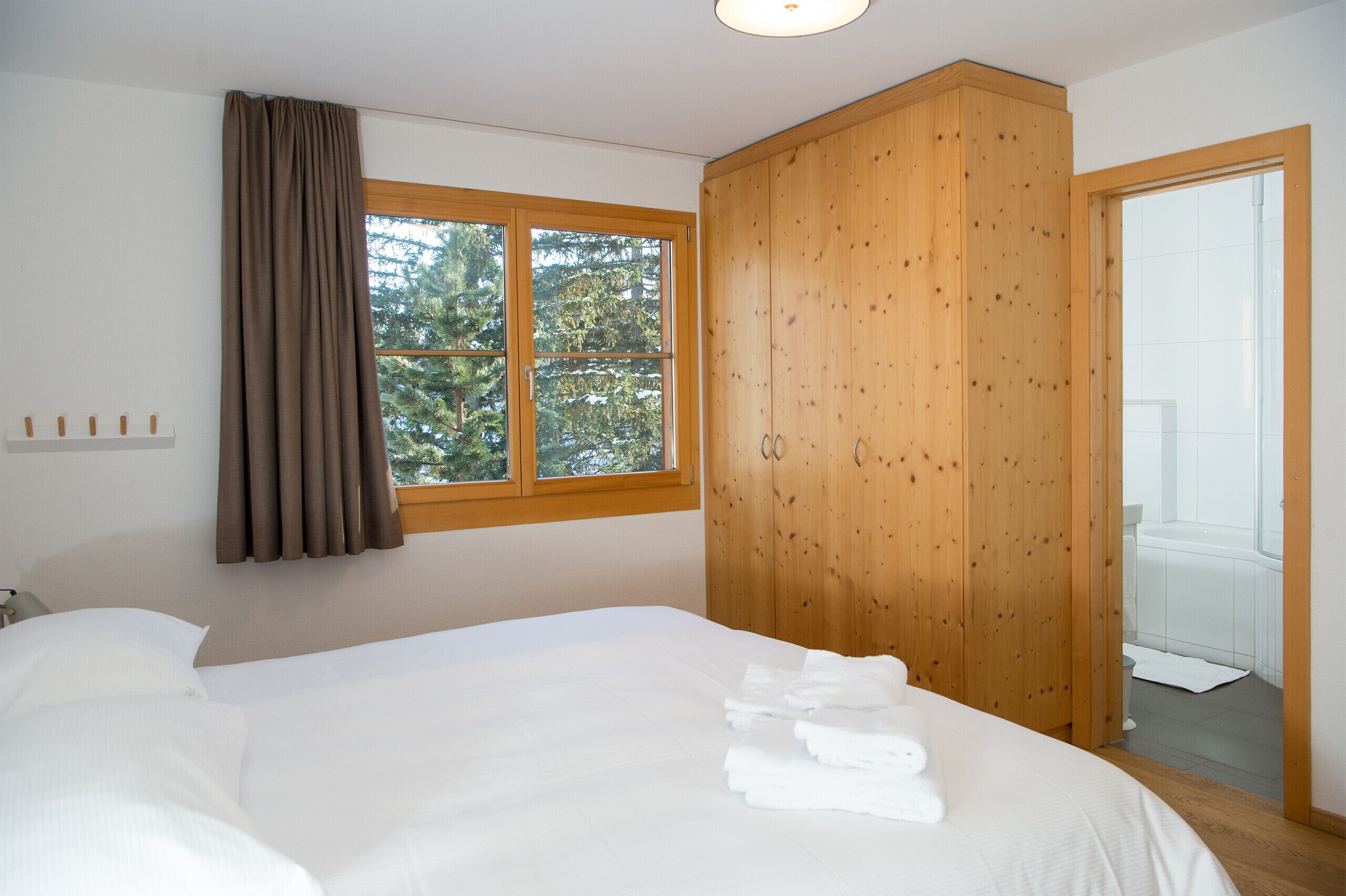 A bedroom with a large bed, a wooden wardrobe, a window with curtains, and a view of trees outside.