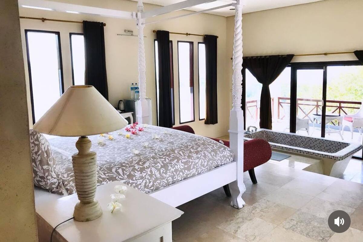 A bedroom with a four-poster bed, a sitting area with a red chair, a bathtub, and a balcony with outdoor furniture.