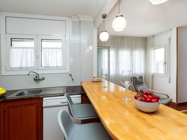 A modern kitchen with a wooden countertop, a built-in dishwasher, and a window with curtains. There are two wine glasses and a bowl of fruit on the counter.