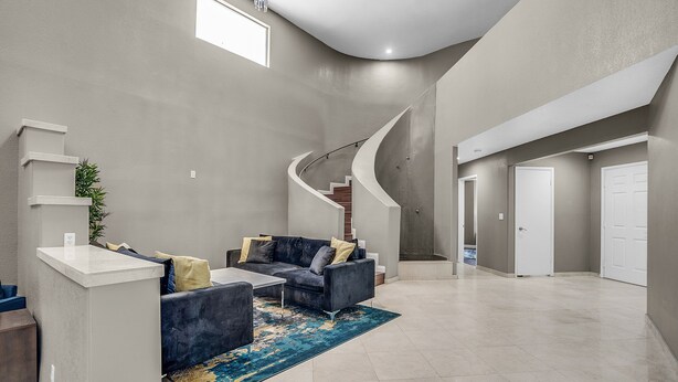 A spacious living area with a curved staircase, a dark grey sofa set, and a blue patterned rug. - Grand Casa (Las Vegas)