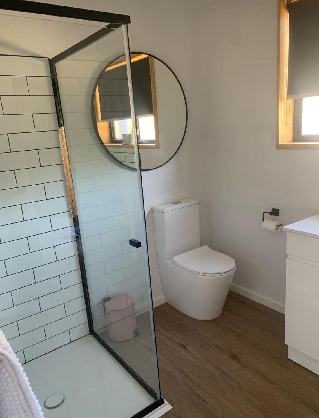 A modern bathroom with a glass shower enclosure, a white toilet, and a round mirror.