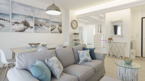 En moderne stue med en grå sofa, et spiseområde med et bord og stole, og et køkken med hvide skabe og et bord med glasplade.