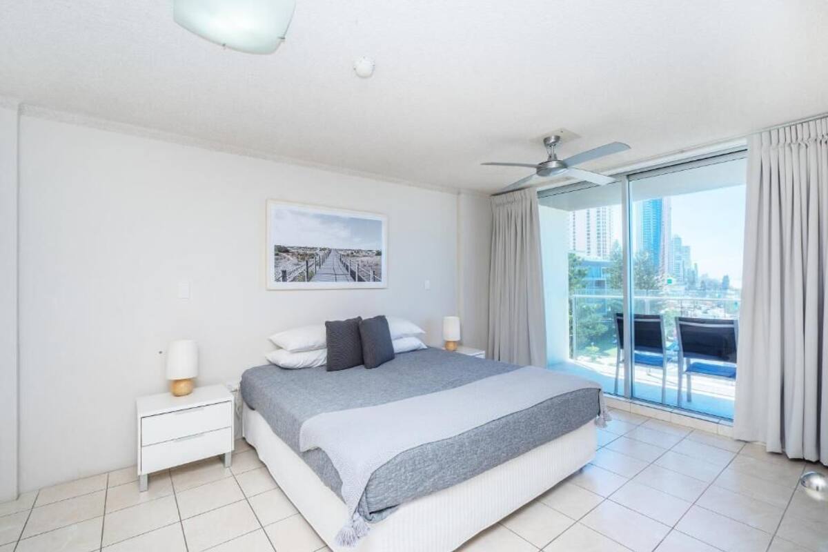 A modern hotel room with a large bed, a ceiling fan, a city view, and a balcony with chairs.