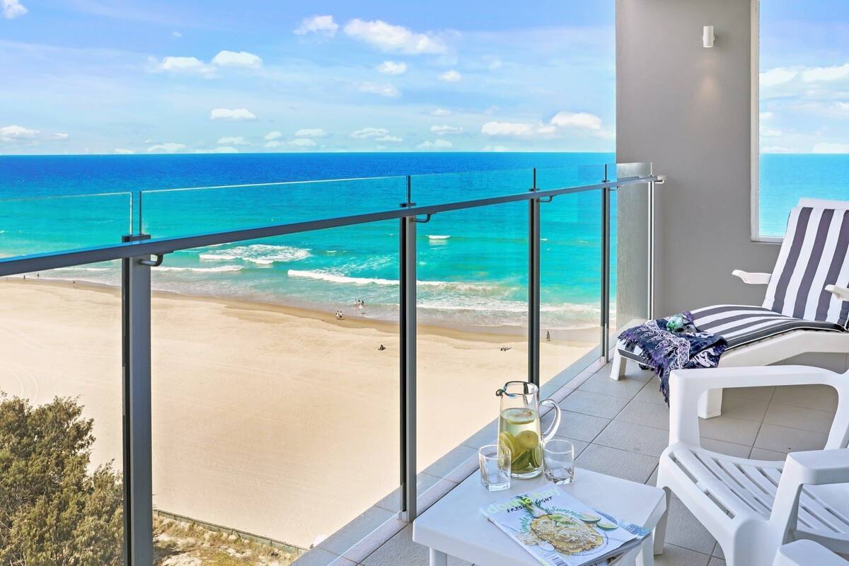 A balcony with a view of a beach and ocean. There is a table with a pitcher of lemonade, a glass, and a magazine.