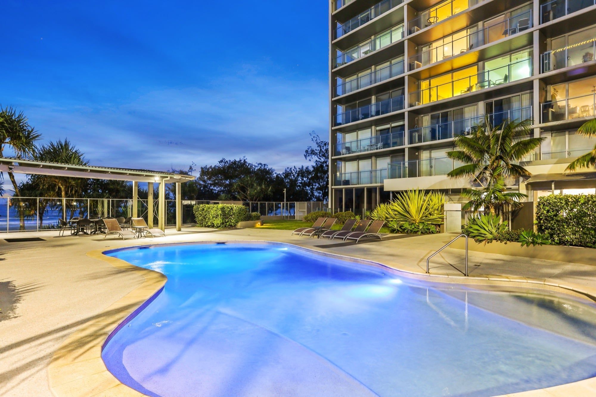 A swimming pool with lounge chairs and a covered seating area, surrounded by a modern building with multiple balconies.