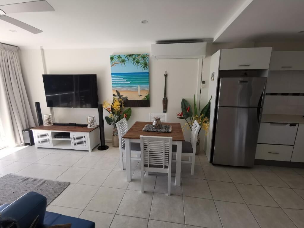 Un salon moderne avec un téléviseur à écran plat, une table à manger en bois avec des chaises blanches, un coin cuisine doté d’un réfrigérateur en acier inoxydable et un tableau sur le mur représentant un paysage de plage.