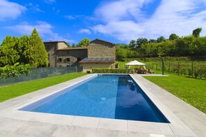 Une piscine rectangulaire bordée d’une terrasse en pierre, entourée d’une pelouse bien entretenue et d’un bâtiment en pierre doté d’un toit en pente.
