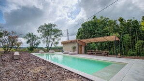 A rectangular swimming pool with a thatched-roof structure and seating area, surrounded by a landscaped garden with trees and a view of a bamboo forest. - Exceptionnel farm house with pool and paddle court (Mareuil en Périgord)