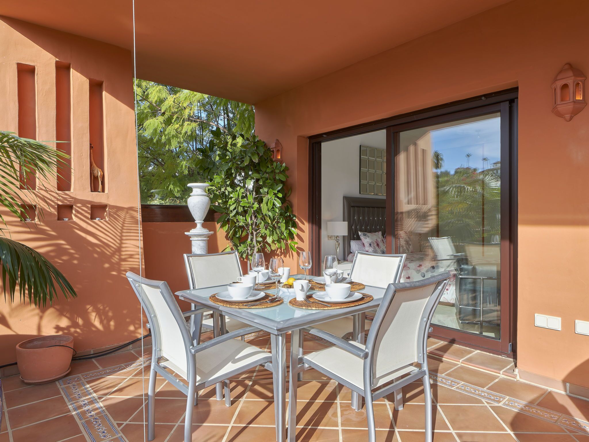 A sunlit patio with a table set for two, surrounded by white chairs, a potted plant, and a large vase.